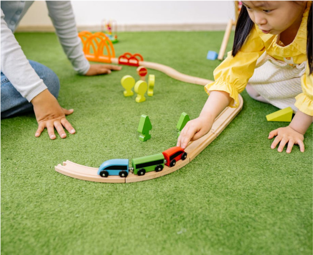 Children playing with toy cars on a wooden train track surrounded by colorful play structures and green grass, illustrating community engagement in toy donation events.