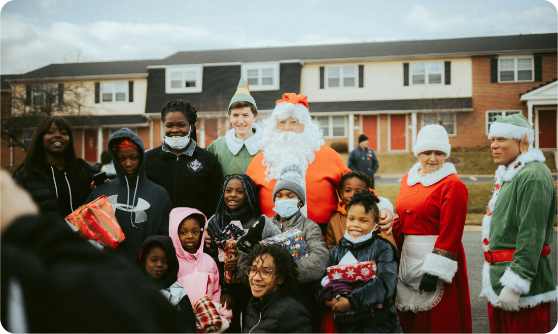 Group of children and volunteers, including Santa Claus and festive characters, smiling while holding wrapped gifts during a holiday toy distribution event organized by GoGo's RePlay Toys.