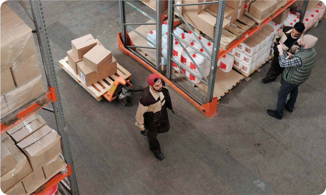 Warehouse volunteers assisting with toy sorting and organization, including a person using a pallet jack to move boxes and two individuals discussing tasks among shelves filled with donations.