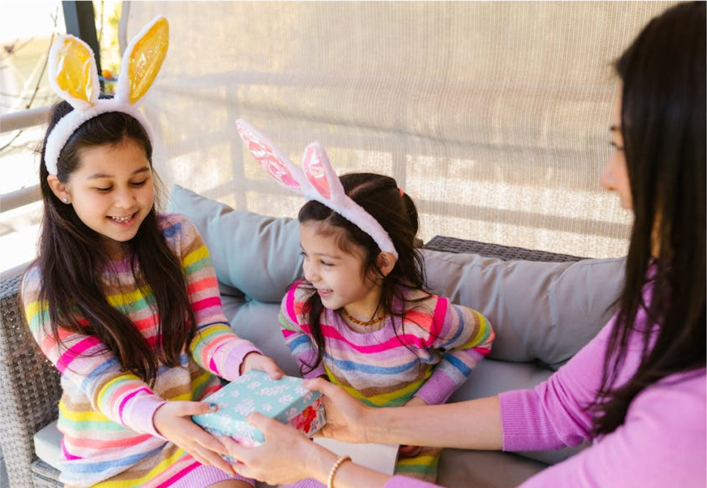 Two girls wearing bunny ears joyfully receiving a wrapped gift from a woman, showcasing the spirit of giving and celebration, relevant to GoGo's RePlay Toys events focused on toy donations for children in need.