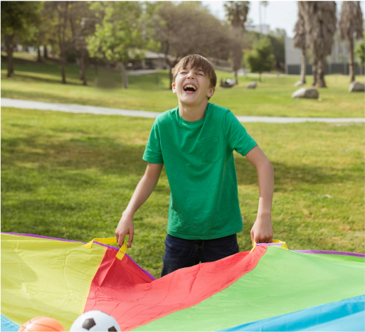 Child joyfully playing with a colorful parachute in a park, surrounded by soccer and beach balls, promoting community engagement and fun at GoGo's RePlay Toys events.