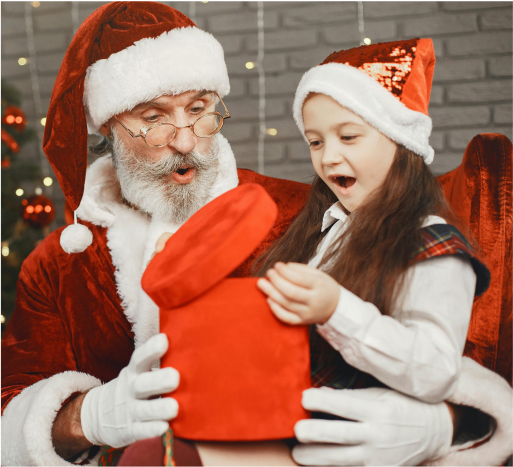 Santa Claus and young girl joyfully opening a red gift box during a festive holiday event, decorated Christmas tree in the background.