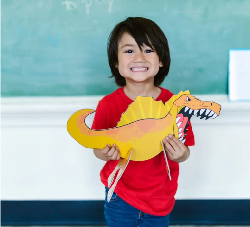 Smiling child holding a colorful dinosaur craft in a classroom setting, representing creativity and playfulness related to community toy events.